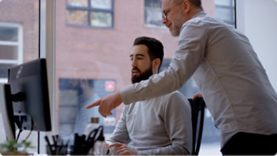 Two people at a desk, one seated at a computer while the other points at the screen.