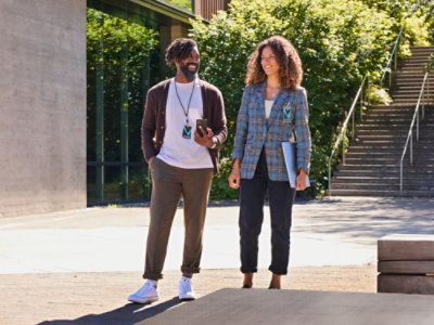 男性と女性が屋外に立っています。女性はチェック柄のジャケットを着ており、男性は電話を持っています。