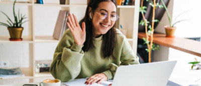 A woman waving at a laptop with a potted plant in the background.