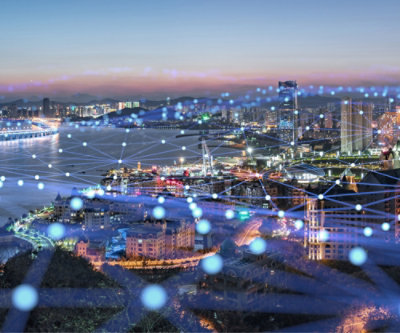 Aerial view of a coastal city at dusk, showcasing illuminated streets and buildings, overlaid with a glowing digital network grid.