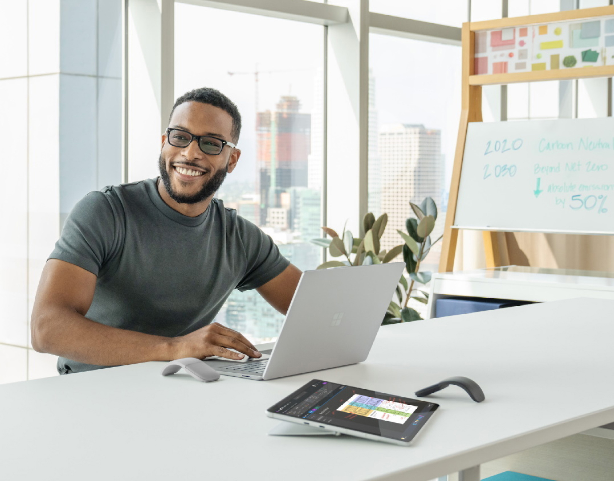 タブレット、電話、プレゼンテーション ボードを備えた最新のオフィス デスクでノート PC を使用している眼鏡をかけて微笑んでいる男性