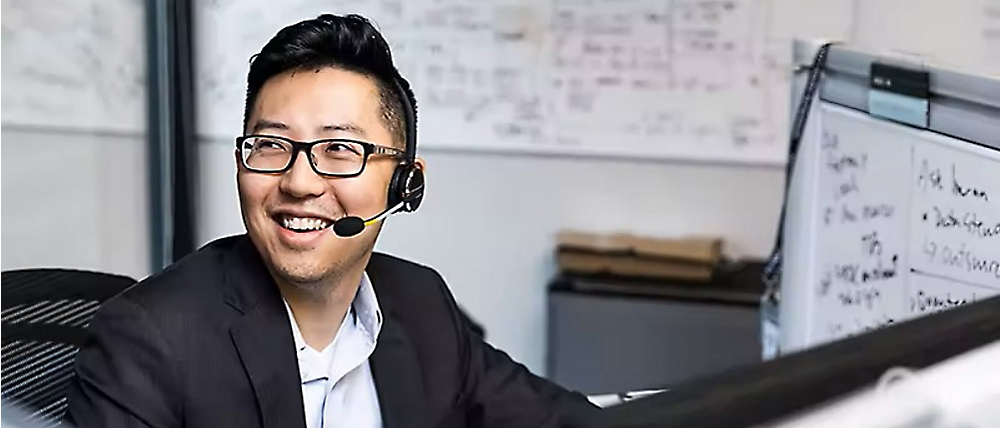 Um homem sorridente, usando fone de ouvido e óculos, olhando para uma tela de computador em um escritório com um quadro de comunicações em segundo plano.