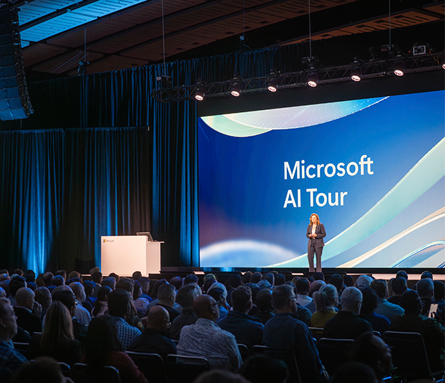 A speaker stands on stage at the Microsoft AI Tour, addressing a large audience. 
