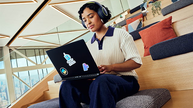 A woman wearing headphones sits on wooden steps in a bright, modern area with large windows. She is using a laptop decorated with colorful stickers. The steps are adorned with cushions in various colors, and a partial city view is visible through the windows. The scene conveys a relaxed, contemporary workspace atmosphere..