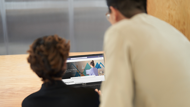 Two people are focused on a laptop screen displaying a website. The website features geometric graphics and a section with text. Both individuals are viewed from behind, suggesting collaboration or shared interest in the content. The setting appears to be a light-colored wooden table in an indoor environment, creating a casual and professional atmosphere.