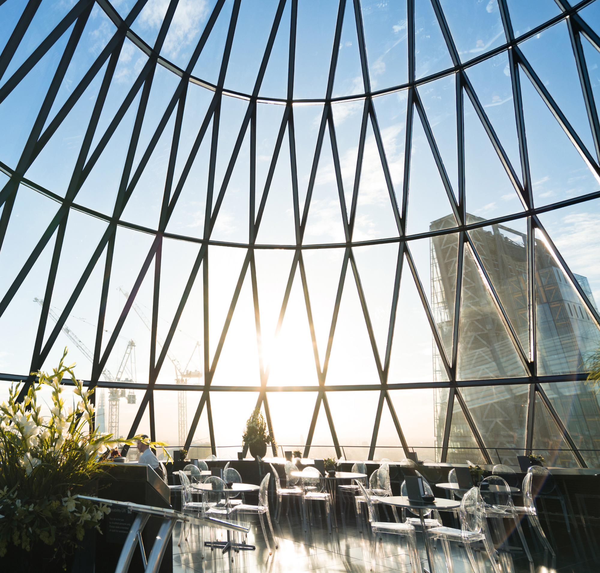 Sunlight streams through a large geometric glass dome overlooking a cityscape, illuminating an interior dining area 
