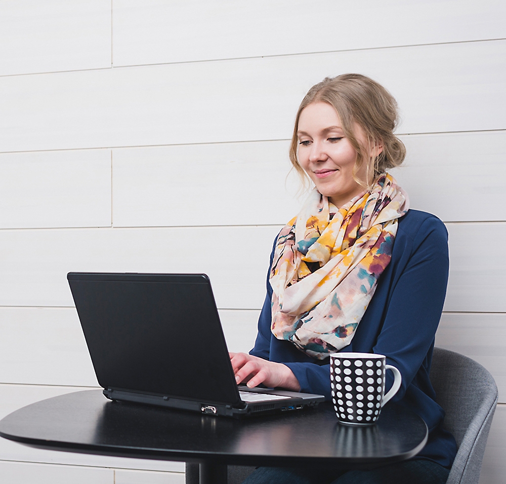 一位身穿蓝色毛衣、戴着彩色围巾的女士在桌子旁用笔记本电脑微笑，旁边放着一个咖啡杯。