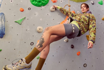 A person in climbing gear ascends an indoor climbing wall, with a rope securing them and various handholds visible.
