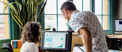 ผู้เชี่ยวชาญสองคนกําลังพูดคุยเกี่ยวกับกราฟบนหน้าจอคอมพิวเตอร์ในสํานักงานที่สว่างไสวพร้อมหน้าต่างขนาดใหญ่และต้นไม้ตกแต่ง