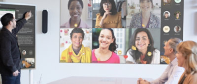 A business meeting with one man presenting to colleagues in a room and others participating virtually on a large screen.