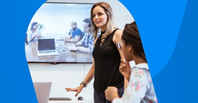 Osoba stoji u sobi za sastanke u kojoj se video poziv prikazuje na velikom ekranu na zidu iza nje