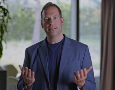 A man wearing a blue suit stands near a brown curtain.