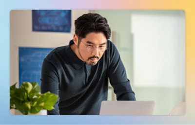 A man wearing glasses and a black shirt is looking at a laptop near a green leafy plant in a white pot.
