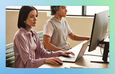 Eine Person tippt an einem Schreibtisch auf einem Laptop, während eine andere Person in der Nähe ein Headset trägt, möglicherweise in einer Besprechung oder während eines Supportgesprächs.