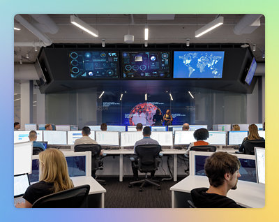 A group of people in a room with computers, with a close-up of a screen and a black office chair.