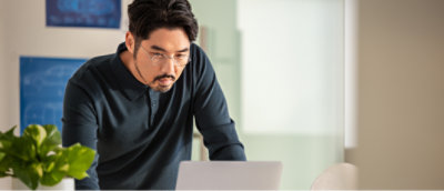 A man in a black shirt looking at a laptop with a blurry green leafy plant in the background.