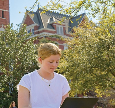 Женщина в белой рубашке сидит на улице и работает за ноутбуком.