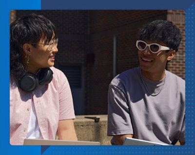 Два человека улыбаются друг другу на улице у кирпичного здания в солнечный день. Один из них в наушниках и очках, другой — с солнцезащитных очках.