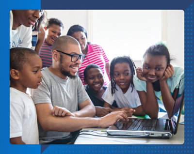 Группа улыбающихся детей разного вида вместе со взрослым собрались у ноутбука, увлеченно общаются и занимаются, демонстрируя чувство общности и радость обучения.