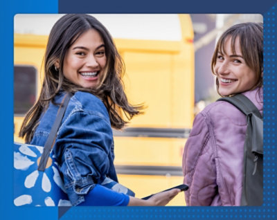 Две женщины идут и улыбаются в камеру. У каждой из них рюкзак, на них обычные куртки, фон ярко-желтого цвета. Радостное настроение.