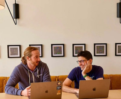 Two men sitting in front of laptops, smiling and looking at each other.