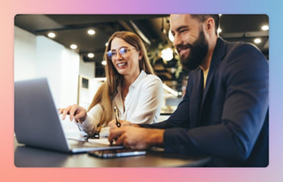 A man and a woman are sitting at a table, looking at a laptop screen, and smiling.