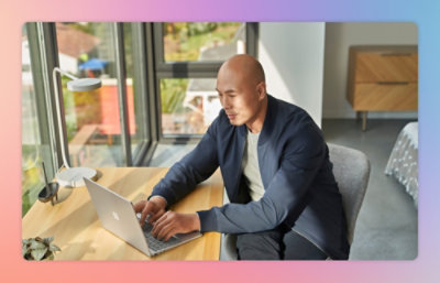 A person sits at a wooden desk typing on a laptop, with sunlight streaming through large windows in the background.
