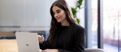 Person sitting at a desk in a modern office, working on a silver laptop with large windows in the background.