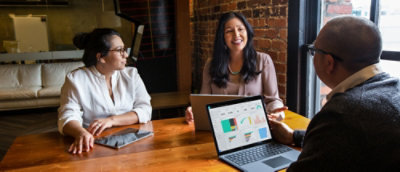 Three people sitting at a wooden table in a modern office, discussing with laptops and a tablet on the table.