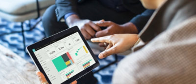 Close-up of a tablet displaying charts and graphs while two people discuss and point at the screen.