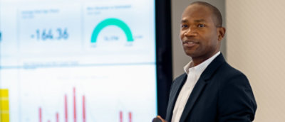 Person standing in front of a large screen displaying charts and a gauge, giving a presentation.
