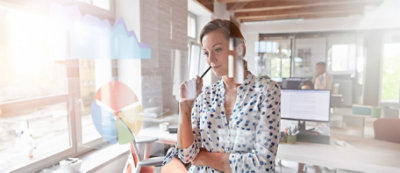 אישה בחולצה עם הדפס עומדת במשרד, מביטה במבט מלא מחשבות, מחזיקה עט ליד שפתיה. 