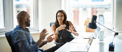 שני עמיתים מנהלים דיון ליד שולחן במשרד מואר היטב, כשברקע מחשב נישא וספל קפה. 