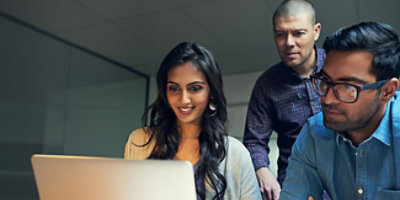 Tri osobe u kancelariji zajedno gledaju u ekran laptop računara. Žena u pravom planu se osmehuje, 