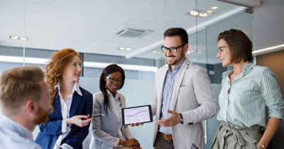 Grupa ljudi u poslovnoj odeći na sastanku u sobi za konferencije. Jedna osoba drži tablet 