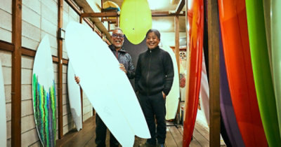 Dvojica nasmejanih muškaraca stoje u radionici, a jedan od njih drži dasku za surfovanje. Oko njih se nalazi još nekoliko dasaka za surfovanje.