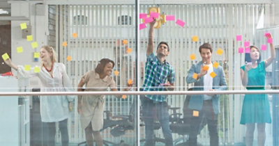 Pet osoba stoji iza staklenog zida prekrivenog raznobojnim lepljivim beleškama, sarađuju i uređuju beleške 