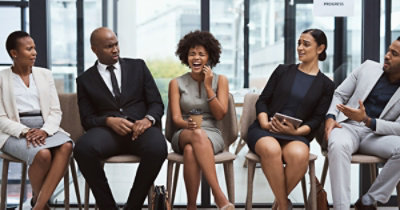 Pet profesionalno odevenih osoba sede u redu stolica i učestvuju u živahnom razgovoru. Žena u sredini se smeje 