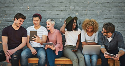 Grupa od šest osoba sedi na klupi postavljenoj uz zid od cigala, komuniciraju i koriste različite elektronske uređaje