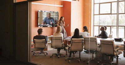 Un gruppo di persone sedute a un tavolo in una sala conferenze guardano una presentazione in un grande schermo a parete.