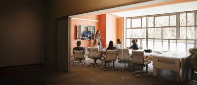 A group of people seated at a table in a conference room watch a presentation on a large wall-mounted screen.