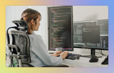 A person in a grey shirt, seated on an office chair, types code on a keyboard in front of two large computer