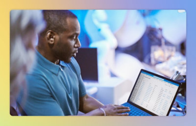 A person in a blue shirt is holding a laptop and showing its screen to another person in a blurred background.