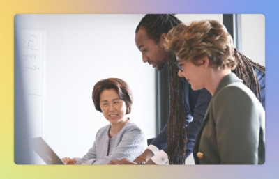 Three professionals, one with dreadlocks, sitting around a laptop in a meeting room, discussing work contentedly.