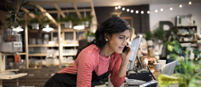 Žena u ružičastoj košulji sa keceljom gleda u ekran računara dok razgovara fiksnim telefonom 