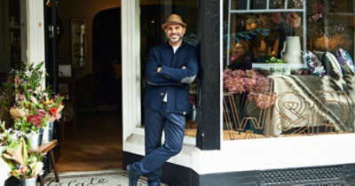 Muškarac sa šeširom stoji skršenih ruku, naslonjen na vrata prodavnice koja su ukrašena cvećem i raznim drugim predmetima 