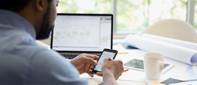 Osoba korzysta ze smartfona, siedząc przy biurku z laptopem, tabletem, kubkiem, dokumentami i długopisem.