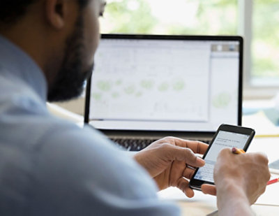 A person sits at a desk in front of a laptop and holds a smartphone. 