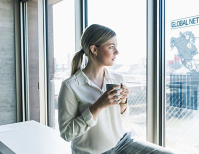 A woman holding a mug looks out a window covered with graphics representing a global network
