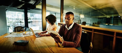 Two individuals are working in an office. One is using a laptop, and the other is holding a tablet.